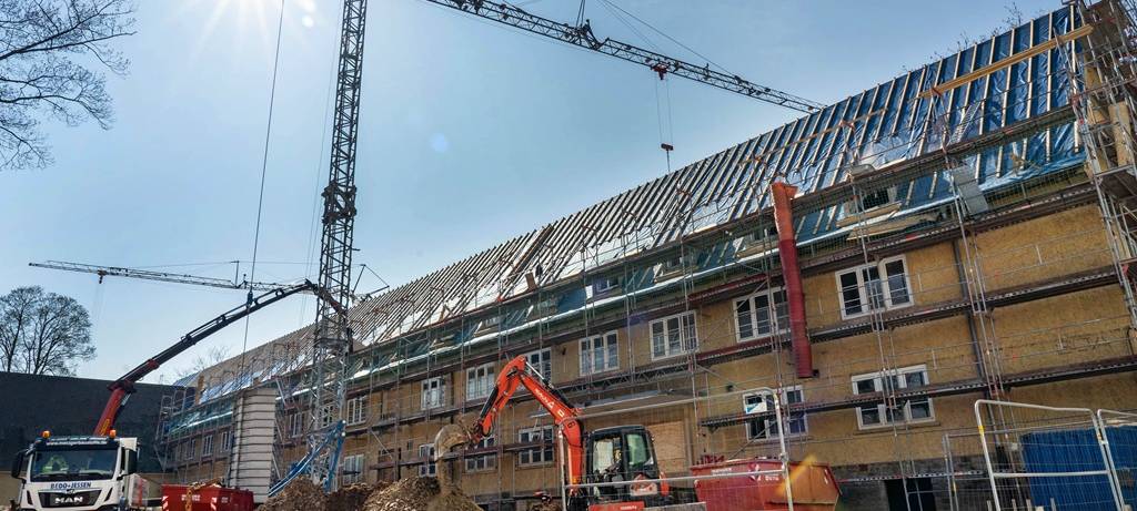 Bild einer Baustelle mit einem langen Kasernengebäude im Gerüst , Kran, Bagger, Aushub und Absperrungen