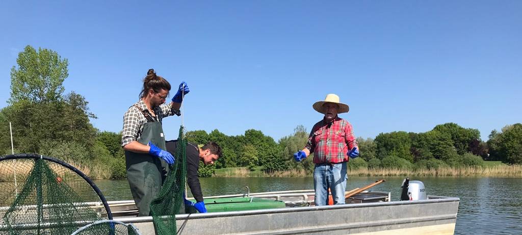 Ein Angelboot aus Stahl mit Außenborder auf dem See. Darauf drei Fischer, zwei jünger, einer davon mit Zopf, einer älter mit Strohhut. Sie setzen Fischreusen ein.